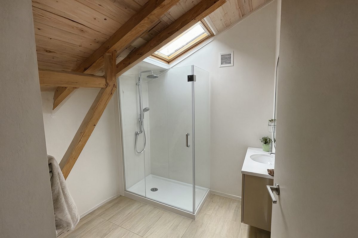 Salle de bain sous combles avec poutres apparentes, fenêtre de toit et cabine de douche